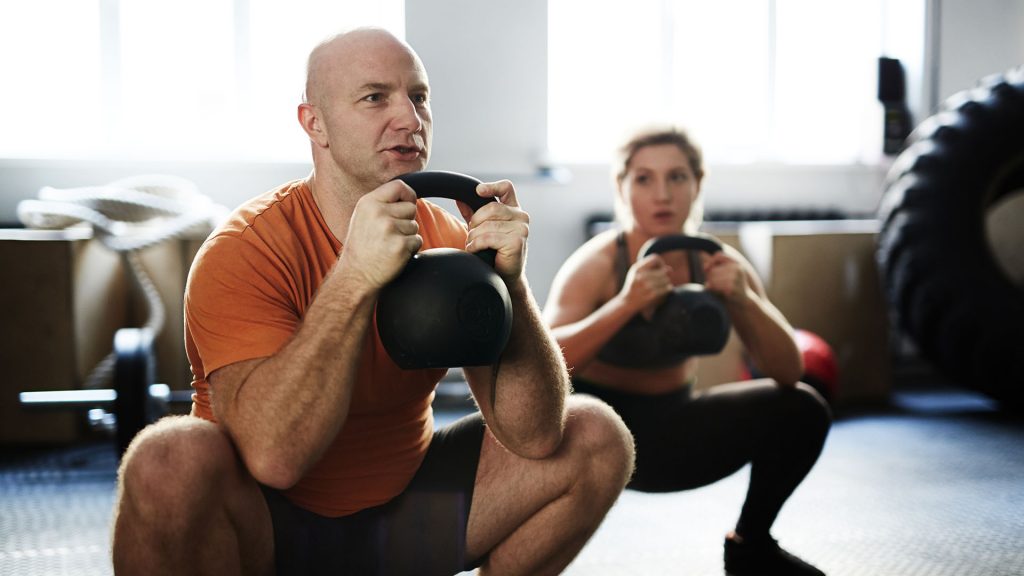 Young sporty woman and middle-aged bodybuilder doing kettlebell squat exercise during intensive training in gym