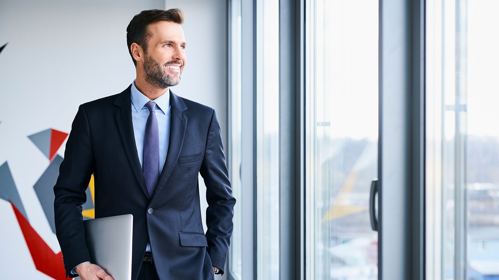 Portrait of middle-aged businessman looking through window while holding laptop